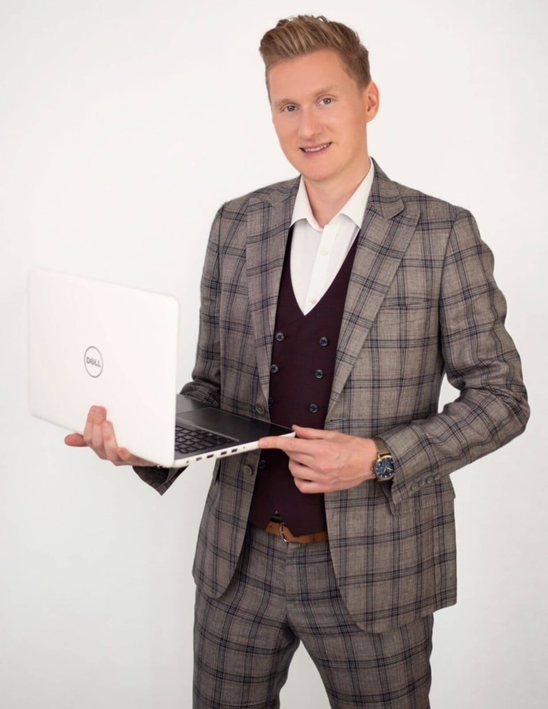 Na zdjęciu widzimy uśmiechniętego, elegancko ubranego Bartłomieja Lewickiego w kratkowanym garniturze w odcieniach szarości z delikatnym brązowym akcentem. Pod marynarką ma ciemną, bordową kamizelkę dwurzędową i białą koszulę. Na lewej ręce nosi stylowy zegarek, a w dłoniach trzyma otwarty biały laptop marki Dell.