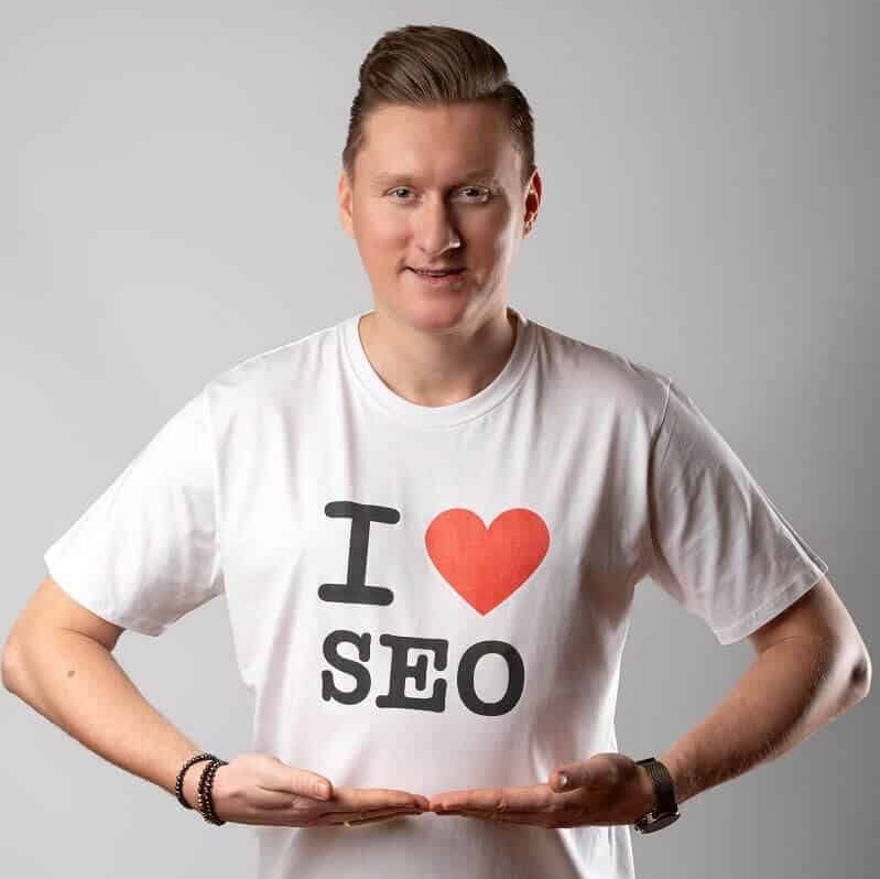 Na zdjęciu widzimy uśmiechniętego Bartłomieja Lewickiego w białej koszulce z napisem ''I ❤️ SEO'', gdzie symbol serca pokazuje słowo ''love''. Jego ręce ułożone są tak, aby podkreślić napis.
