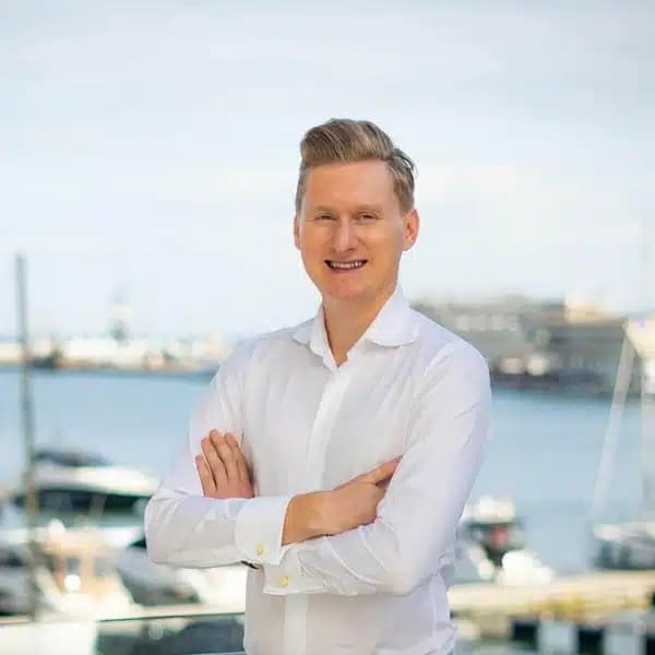Na zdjęciu widzimy uśmiechniętego mężczyznę, który ubrany jest w elegancką białą koszulę. Mężczyzna stoi z założonymi rękami na tle portu.