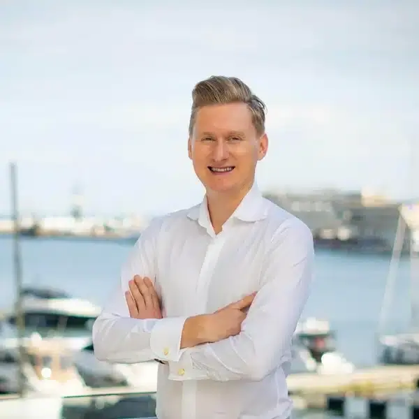 Na pierwszym planie stoi elegancko ubrany mężczyzna w białej koszuli, z rękami skrzyżowanymi na piersi. W tle rozciąga się marina pełna łodzi i żaglówek, a za nią delikatnie zarysowane budynki tworzące spokojną, nadmorską atmosferę. Kontrast między formalnym strojem a wakacyjnym pejzażem podkreśla ciekawe zestawienie stylu i miejsca.