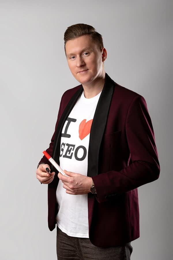 Na zdjęciu widać mężczyznę, który jest portretowany. Ma zaczesane do góry jasne włosy. Ubrany jest w elegancką, bordową marynarkę z czarnymi, połyskującymi klapami, pod którą ma białą koszulkę z napisem "I ❤ SEO". Na lewym nadgarstku widać złoty zegarek. W prawej ręce trzyma czerwono-biały marker. Mężczyzna patrzy wprost w obiektyw, ma delikatny uśmiech na twarzy. Tło jest jednolite, jasnoszare.