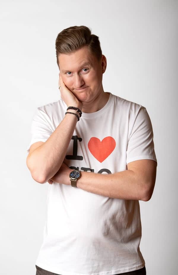 Na zdjęciu jest uśmiechnięty mężczyzna, który ma na sobie białą koszulkę z dużym czerwonym sercem i napisem "I ❤ SEO". Mężczyzna podpiera jedną ręką policzek, a drugą ma złożoną na piersi. Na nadgarstkach ma zegarek i bransoletki. Patrzy wprost w obiektyw, a tło jest jednolite i jasne.