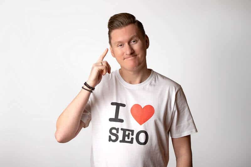 Na zdjęciu jest mężczyzna, który ma na sobie białą koszulkę z dużym napisem "I ❤ SEO". Ma podniesiony palec wskazujący do skroni, jakby o czymś intensywnie myślał. Mężczyzna ma krótkie, jasne włosy zaczesane do góry, a na nadgarstku nosi bransoletkę z ciemnych koralików. W tle widać jednolitą, jasną ścianę.