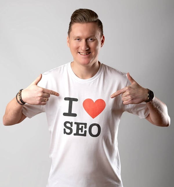 Na zdjęciu widzimy mężczyznę ubranego w białą koszulkę z napisem „I ❤️ SEO”, gdzie czerwone serce symbolizuje zamiłowanie do optymalizacji stron internetowych. Mężczyzna uśmiecha się i wskazuje obiema rękami na nadruk, wyraźnie podkreślając swój entuzjazm wobec SEO. Tło jest jasnoszare i jednolite, co sprawia, że cała uwaga skupia się na przekazie koszulki i pozytywnej energii osoby. Grafika ma lekki, nowoczesny charakter i świetnie nadaje się do promocji usług związanych z marketingiem internetowym.