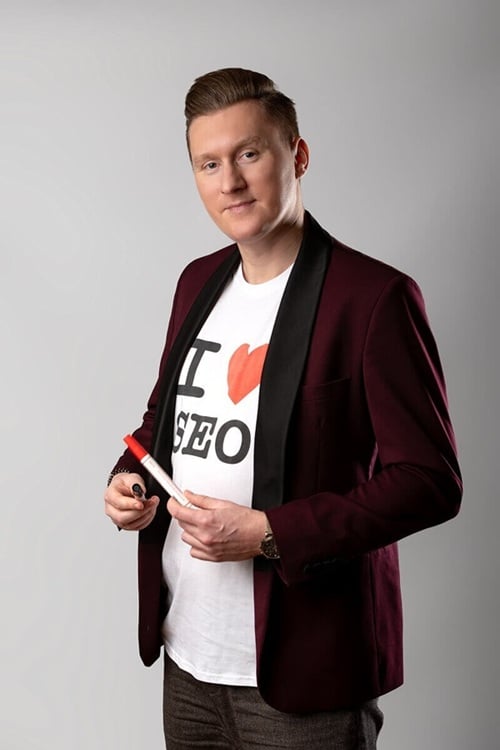 Na zdjęciu widzimy osobę ubraną w elegancką bordową marynarkę z czarnymi klapami, pod którą znajduje się biała koszulka z wyraźnym napisem „I ♥ SEO”. Czerwone serce symbolizuje zamiłowanie do optymalizacji stron internetowych. Osoba trzyma w dłoniach czerwono-biały marker i patrzy prosto w obiektyw z lekkim uśmiechem, co nadaje zdjęciu profesjonalny, ale jednocześnie swobodny charakter. Tło jest jasnoszare i jednolite.