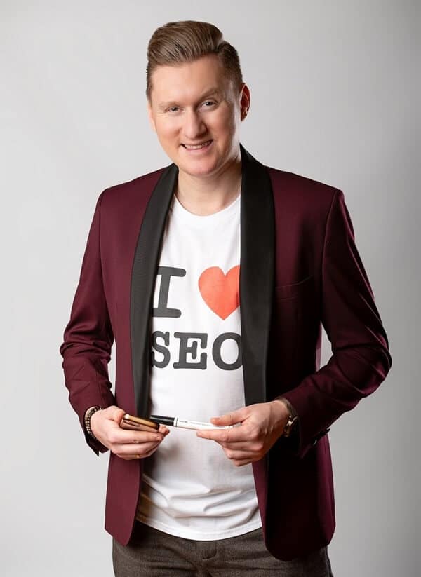Na zdjęciu widzimy uśmiechniętego mężczyznę ubranego w bordową marynarkę z czarnymi klapami, pod którą ma białą koszulkę z napisem „I ❤️ SEO”. W dłoniach trzyma dwa markery, co może sugerować, że zajmuje się edukacją lub prezentacjami związanymi z tematyką SEO.