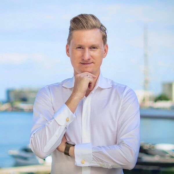 Na zdjęciu widzimy elegancko ubranego mężczyznę stojącego na tle nadmorskiego krajobrazu. Ma na sobie białą koszulę z mankietami ozdobionymi złotymi spinkami oraz zegarek na nadgarstku, co podkreśla jego profesjonalny wizerunek. Jego fryzura jest starannie ułożona, a poza – z ręką opartą o brodę – sugeruje pewność siebie i refleksję.