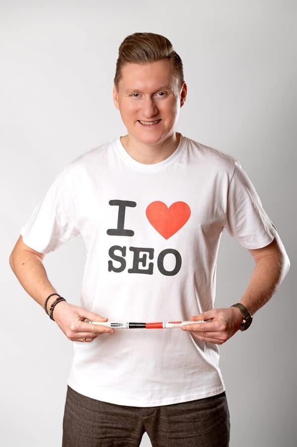 Na zdjęciu widzimy uśmiechniętego mężczyznę w białej koszulce z wyraźnym napisem „I ❤️ SEO”, który symbolizuje jego pasję do optymalizacji stron internetowych. Trzyma w dłoniach dwa markery – czerwony i czarny – ułożone poziomo, co kolorystycznie nawiązuje do nadruku na koszulce i tworzy spójną, przemyślaną kompozycję. Jego stylizacja jest prosta, ale wyrazista: neutralne tło, schludna fryzura, zegarek i bransoletka dodają osobistego charakteru.