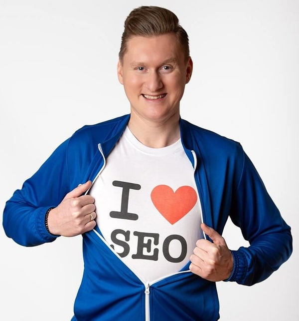 Na zdjęciu widoczny jest uśmiechnięty mężczyzna w niebieskiej bluzie, którą rozsuwa, odsłaniając koszulkę z napisem „I ♥ SEO”. Gest ten podkreśla jego pasję do pozycjonowania stron i marketingu internetowego.